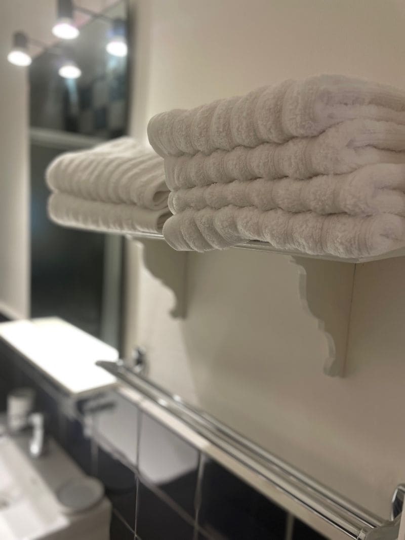 A bathroom shelf displays neatly stacked white towels, providing a fresh and tidy appearance. Reflections can be seen in the mirror above the shelf, and the soft lighting enhances the overall ambiance.