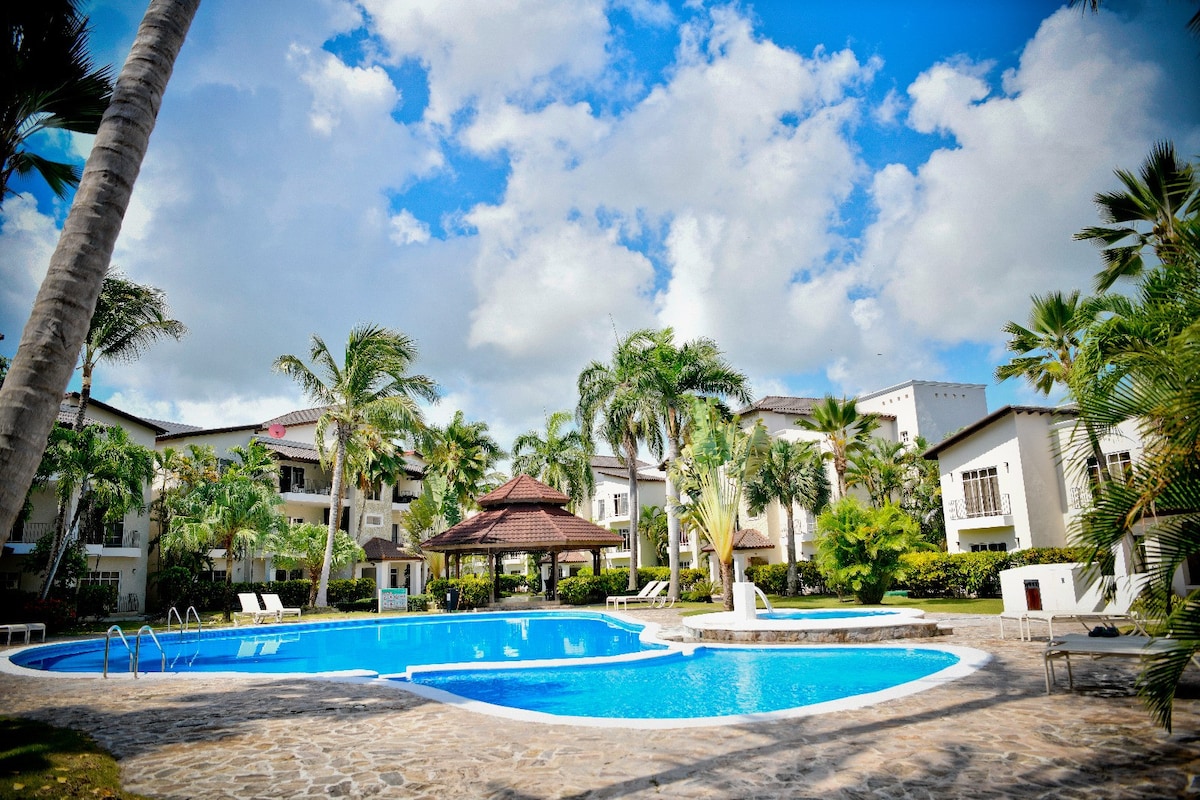 A serene pool area is surrounded by lush palm trees and well-maintained landscaping. The pool features a spacious layout with a separate jacuzzi, complemented by a gazebo. Sun loungers are positioned near the water, inviting relaxation in a tranquil setting.