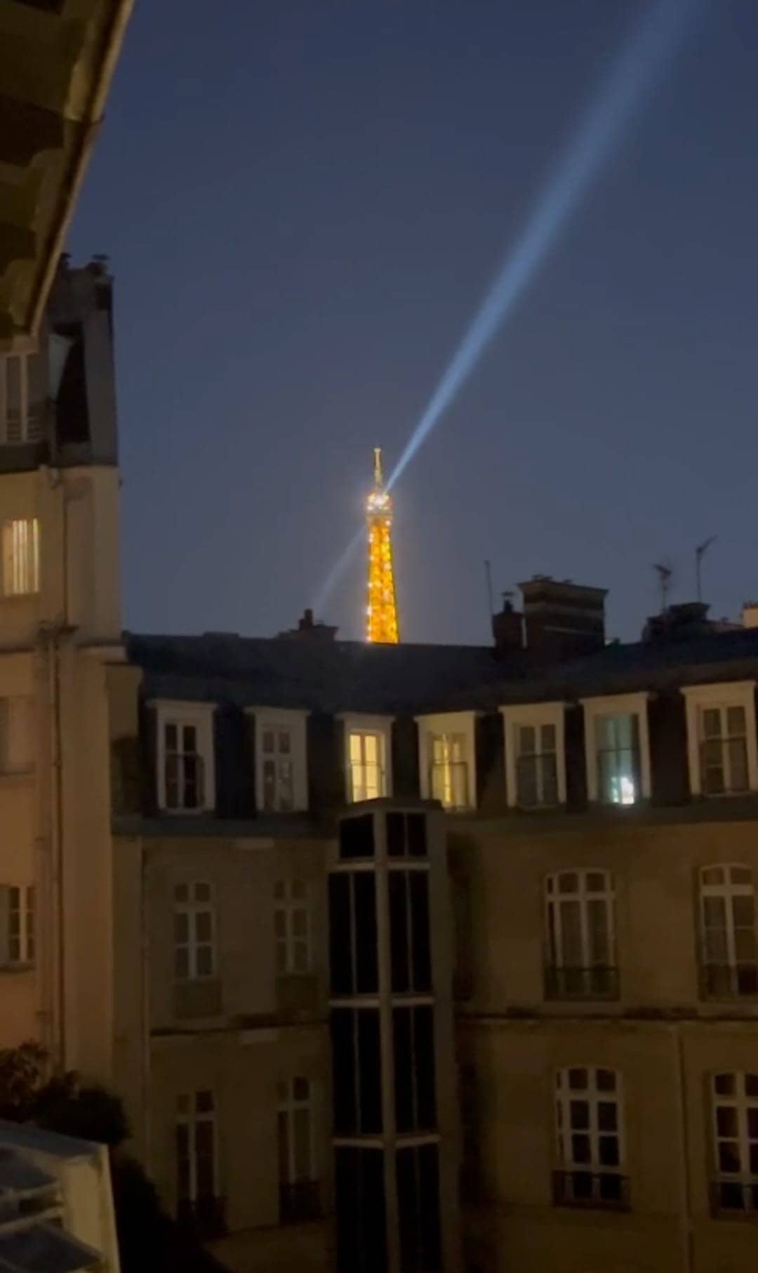 The image captures a nighttime view of the Eiffel Tower, illuminated and showcasing a beam of light radiating from its top. Surrounding buildings feature varying window designs, contributing to the Parisian ambiance.