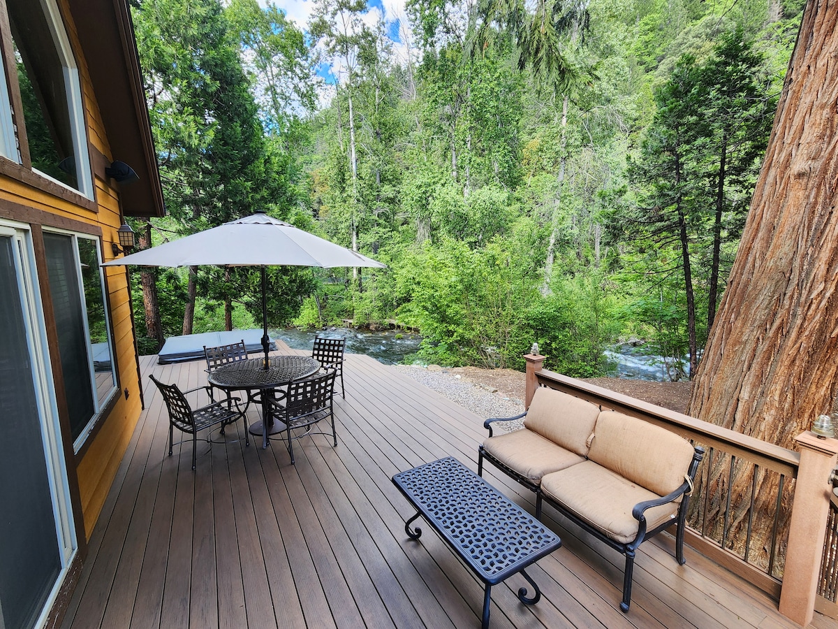 A spacious back deck is displayed, featuring a round dining table with six chairs and a comfortable seating area. An umbrella provides shade, while lush greenery and a flowing river are visible in the background, enhancing the outdoor setting.