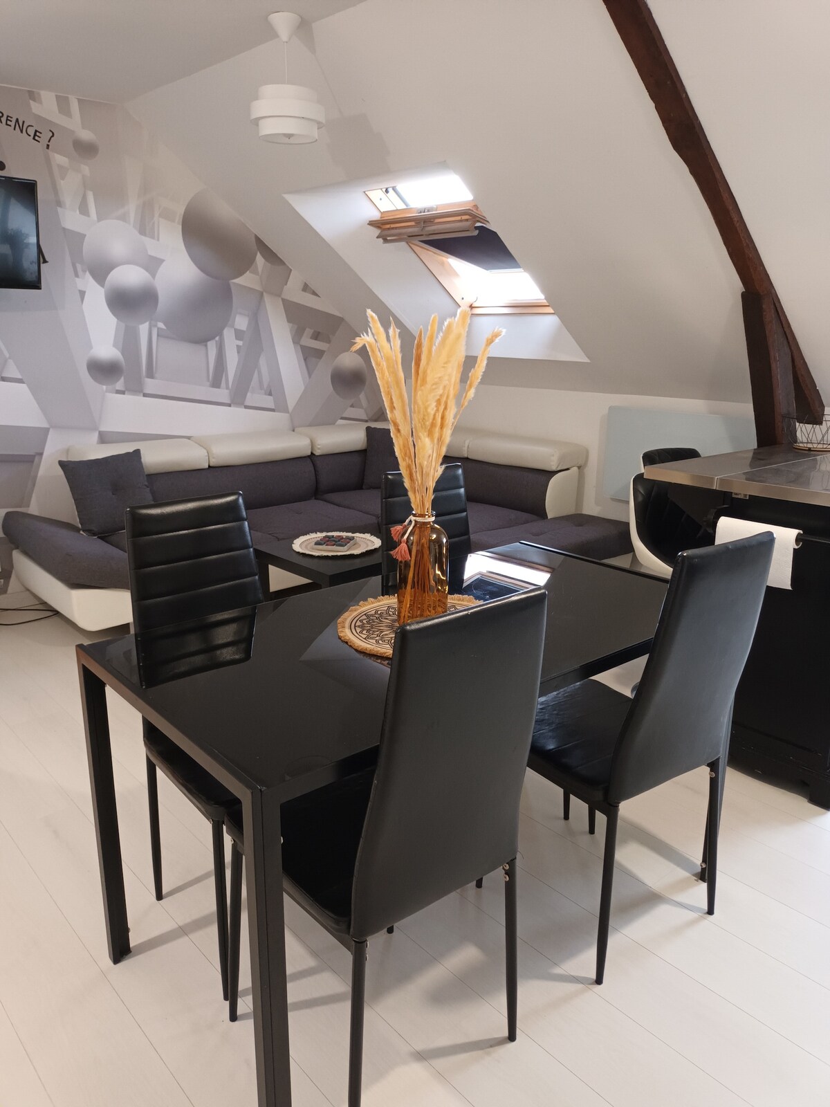 A dining area is featured with a sleek black table surrounded by four black chairs. The space is illuminated by natural light streaming through a skylight, highlighting the modern design. A cozy living area with a sectional sofa is visible in the background, enhancing comfort.