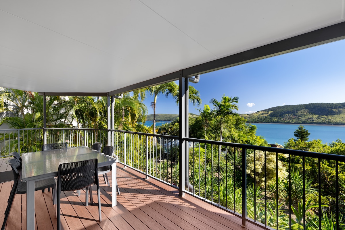 A spacious balcony is provided, featuring a dining table and chairs under a roof. Lush greenery and palm trees frame the view of the sparkling ocean and distant hills, offering a scenic backdrop for outdoor dining.