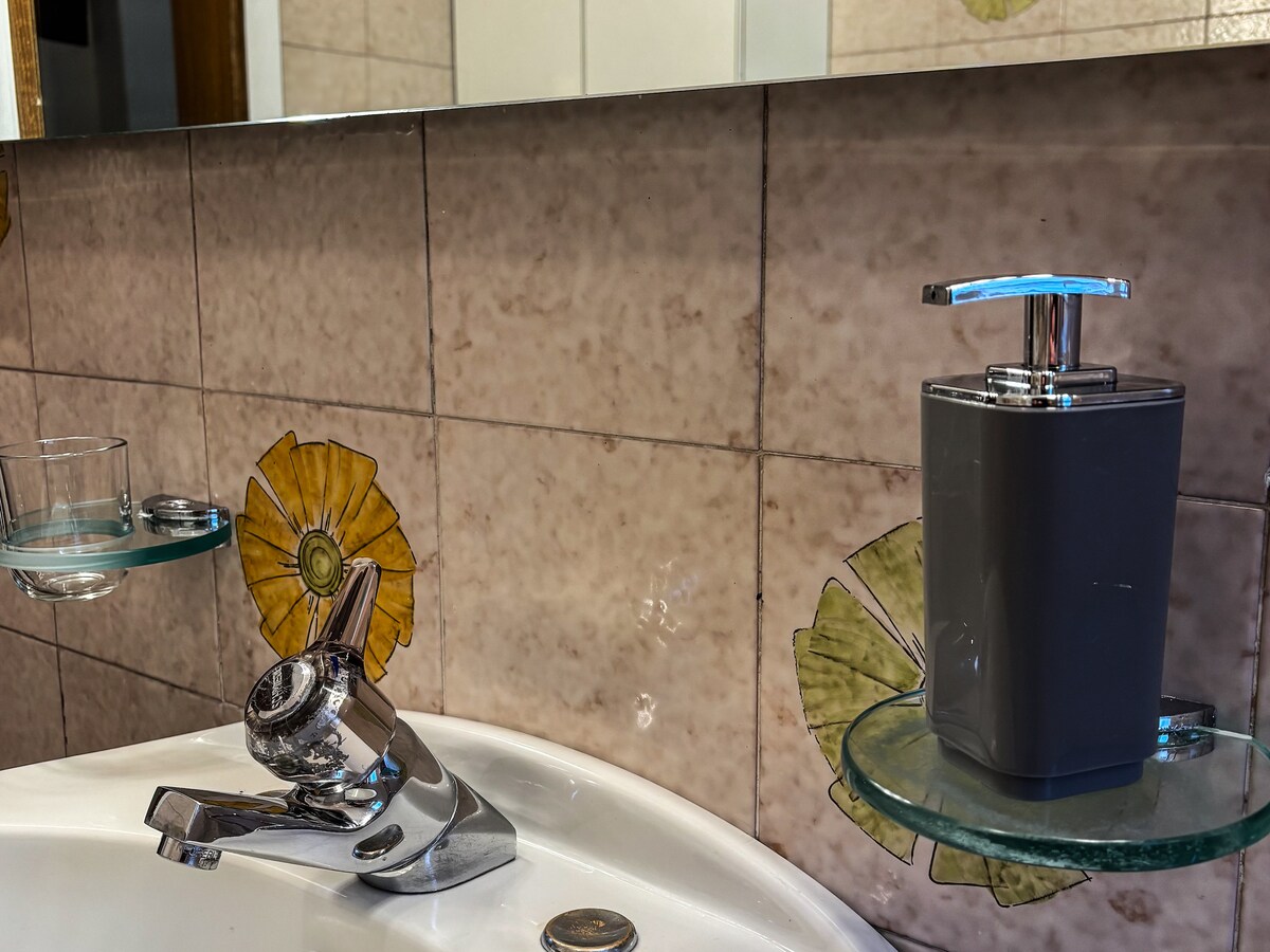 A bathroom sink area is depicted, featuring a modern faucet and two glass shelves. One shelf holds a gray soap dispenser, while the other displays a small glass. Decorative tiles with a floral design enhance the space's character.
