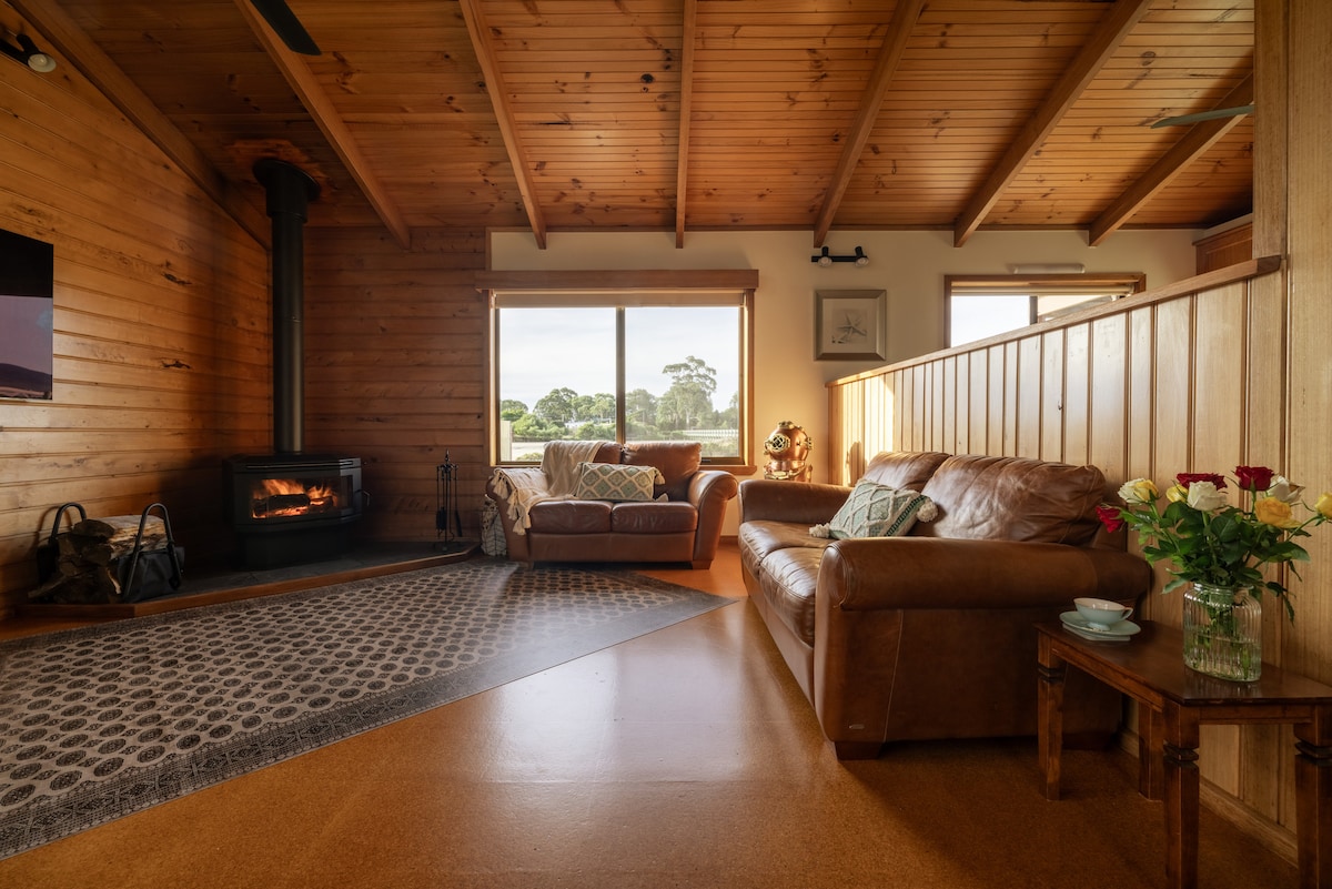 A cozy living area features wooden walls and high ceilings with exposed beams. A leather couch is positioned near a window offering views of greenery outside, while a wood-burning stove adds warmth to the space. A small table holds a vase of fresh roses.