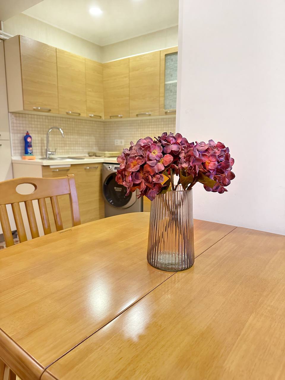 A dining table with a wooden finish features a glass vase containing dried flowers. In the background, an equipped kitchen is visible, showcasing cabinetry and essential appliances. The setting combines inviting natural materials and a functional layout, suitable for dining and gatherings.