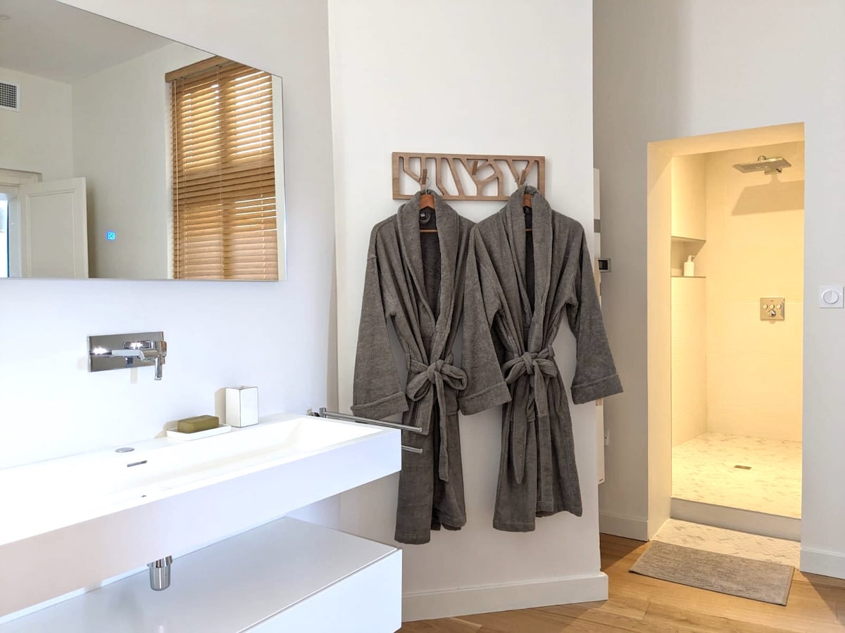 The bathroom features a spacious vanity with a large mirror above it, complemented by two plush bathrobes hanging nearby. An entrance to a shower area can be seen in the background, showcasing a modern and clean aesthetic.