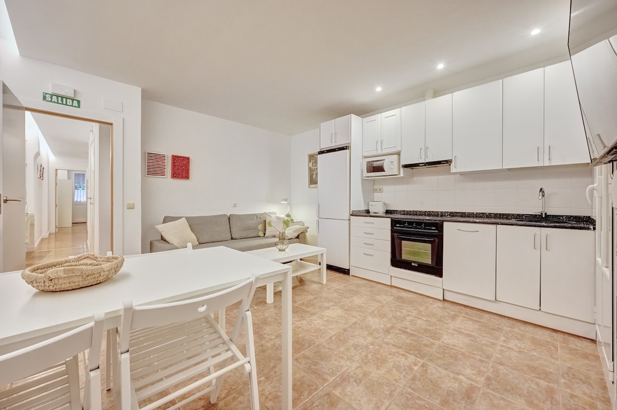 A functional kitchen area features white cabinetry and appliances, including a microwave and oven. A grey sofa sits adjacent to a dining table with white chairs. Natural light brightens the tiled floor and walls, creating an open and airy space.