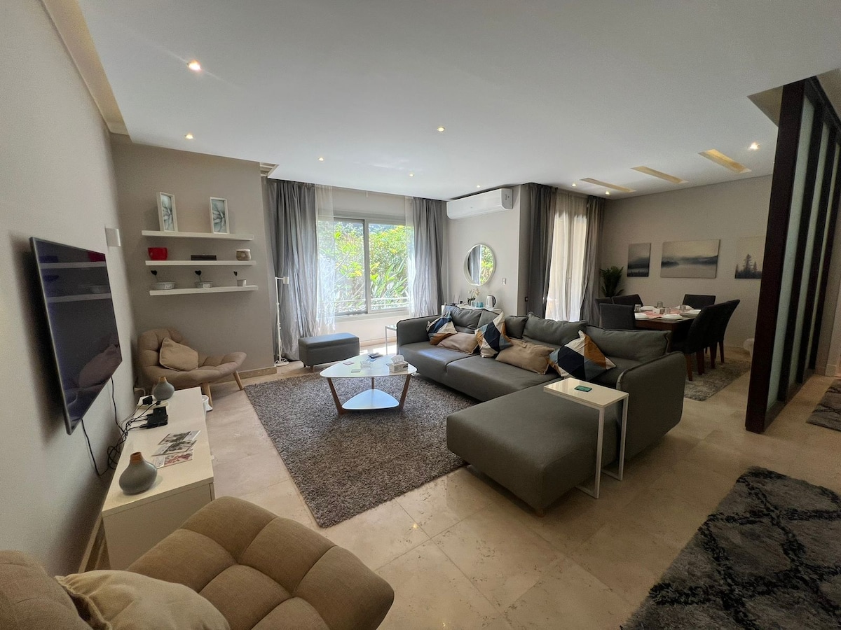 A spacious living area is furnished with a plush sectional sofa and a modern coffee table. Natural light filters through large windows, highlighting the serene decor. A dining area with a round table is visible in the background, accompanied by tasteful wall art and soft drapes.