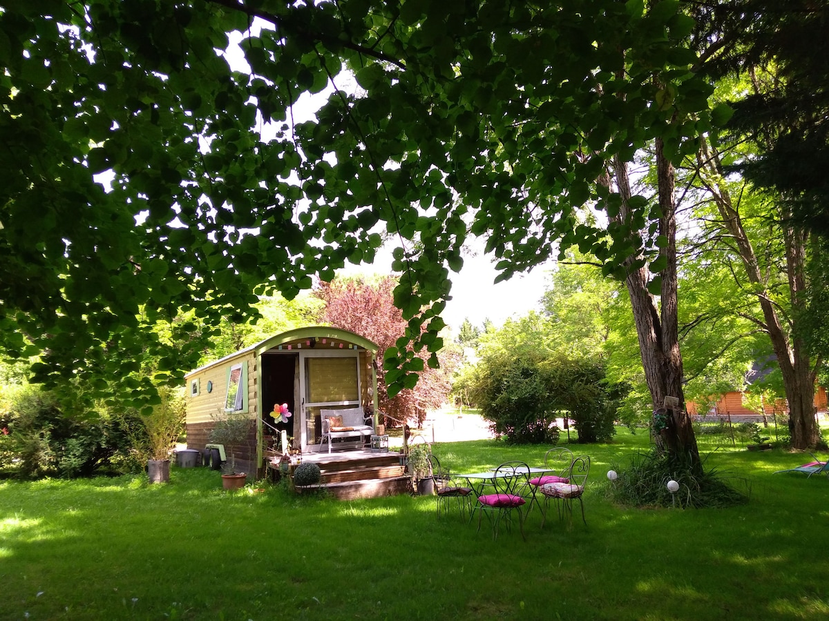 A charming roulotte is nestled under lush green foliage, surrounded by vibrant plants. The entrance features steps leading up to a welcoming porch. A cozy seating area with several garden chairs invites relaxation amidst the spacious grassy area, creating a serene outdoor retreat.