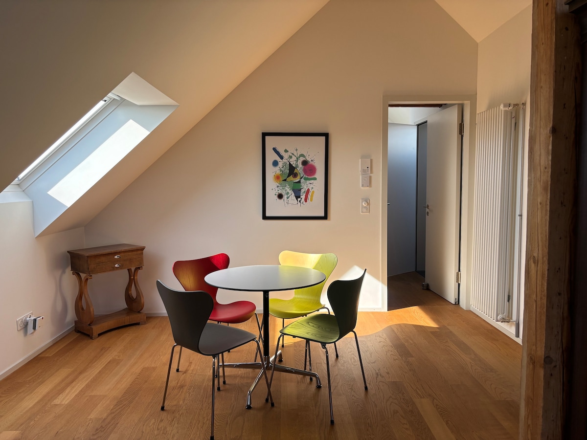 A dining area is presented with a round table surrounded by four colorful chairs in yellow, green, red, and gray. A vibrant artwork adorns the wall, and natural light enters through a skylight, enhancing the spacious feel of the room.