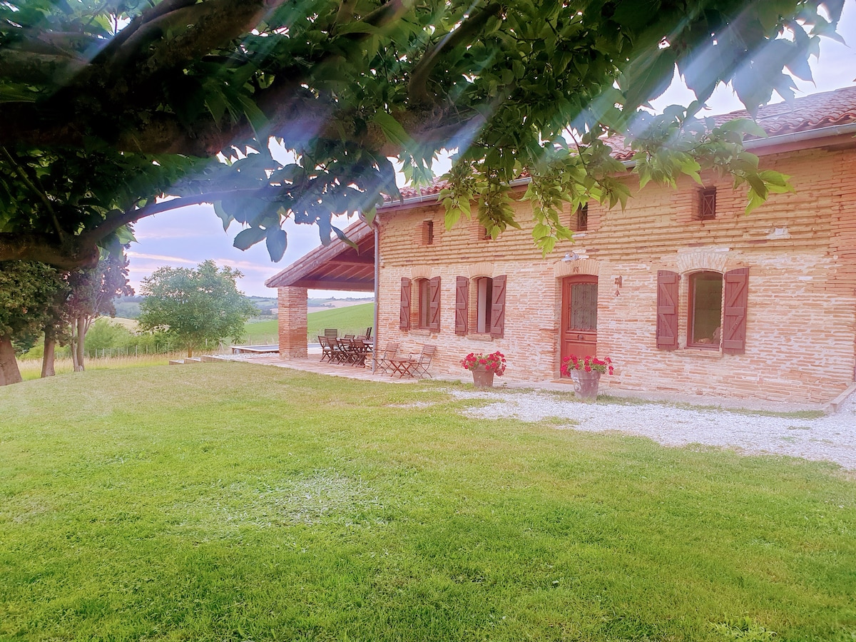 The charming brick farmhouse is framed by lush greenery and a vast expanse of lawn. Several windows create an inviting aspect, while flower pots add touches of color. A shaded terrace is visible, offering space for outdoor dining or relaxation.