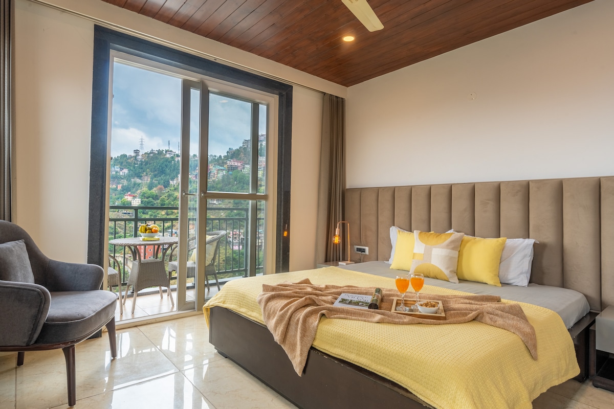 A spacious bedroom features a large bed with yellow accents and a cozy blanket. A comfortable armchair is placed nearby, and a balcony offers scenic views of the surrounding landscape. Natural light floods the room through glass doors, enhancing the inviting atmosphere.