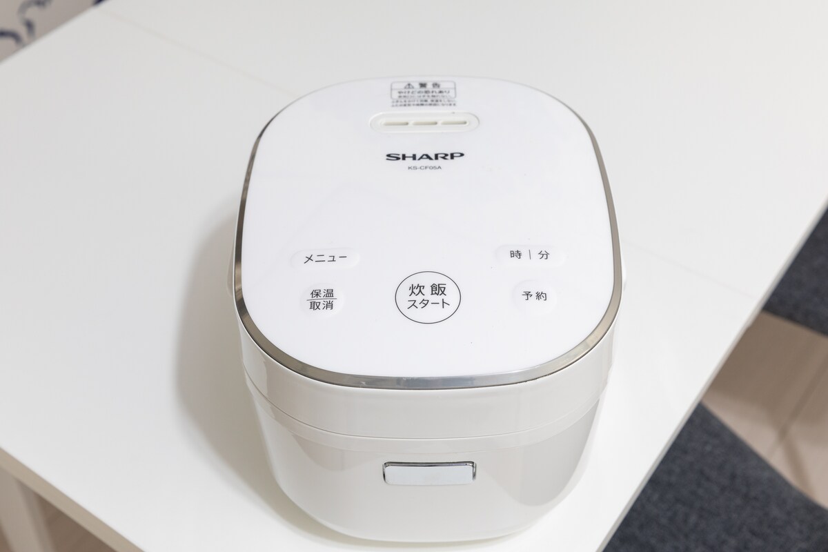 A rice cooker is displayed on a white table. The cooker features a sleek, modern design with a smooth, rounded top and clearly labeled buttons for menu options, timer, and start functions. The overall appearance is clean and well-maintained.