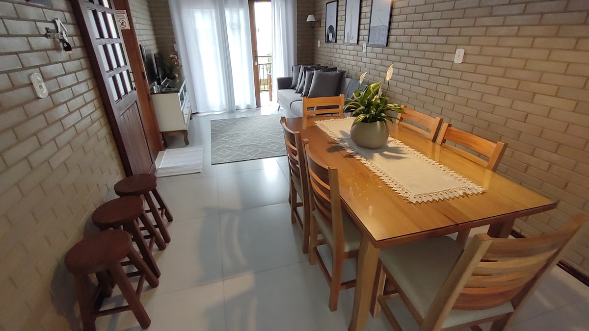 A dining area is visible with a wooden table surrounded by six chairs, while three wooden stools are positioned nearby. Natural light enters through large glass doors leading to a balcony. A comfortable gray sofa is softly illuminated, complementing the brick walls.