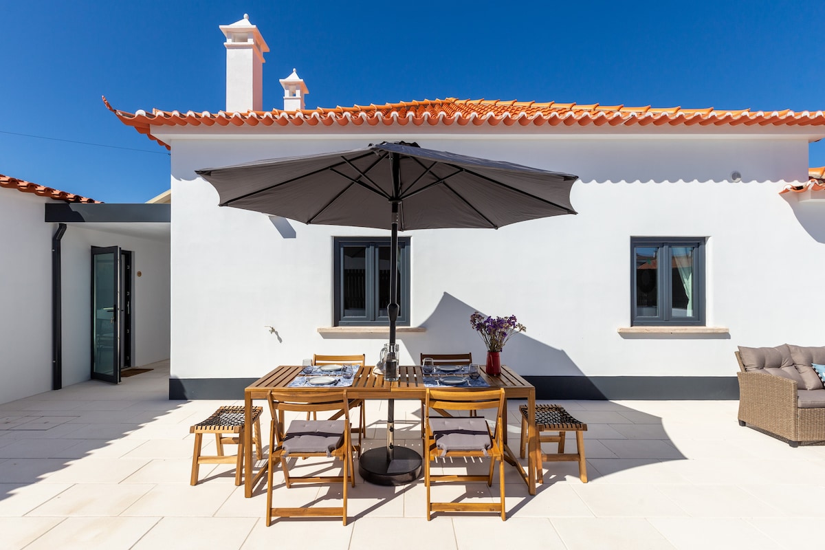 An outdoor dining area is featured under a large umbrella, surrounded by a wooden table and chairs. The patio is adorned with sunlight, and a cozy seating arrangement is visible on one side. The walls of the house are painted in a bright white, contrasting with the terracotta roof.