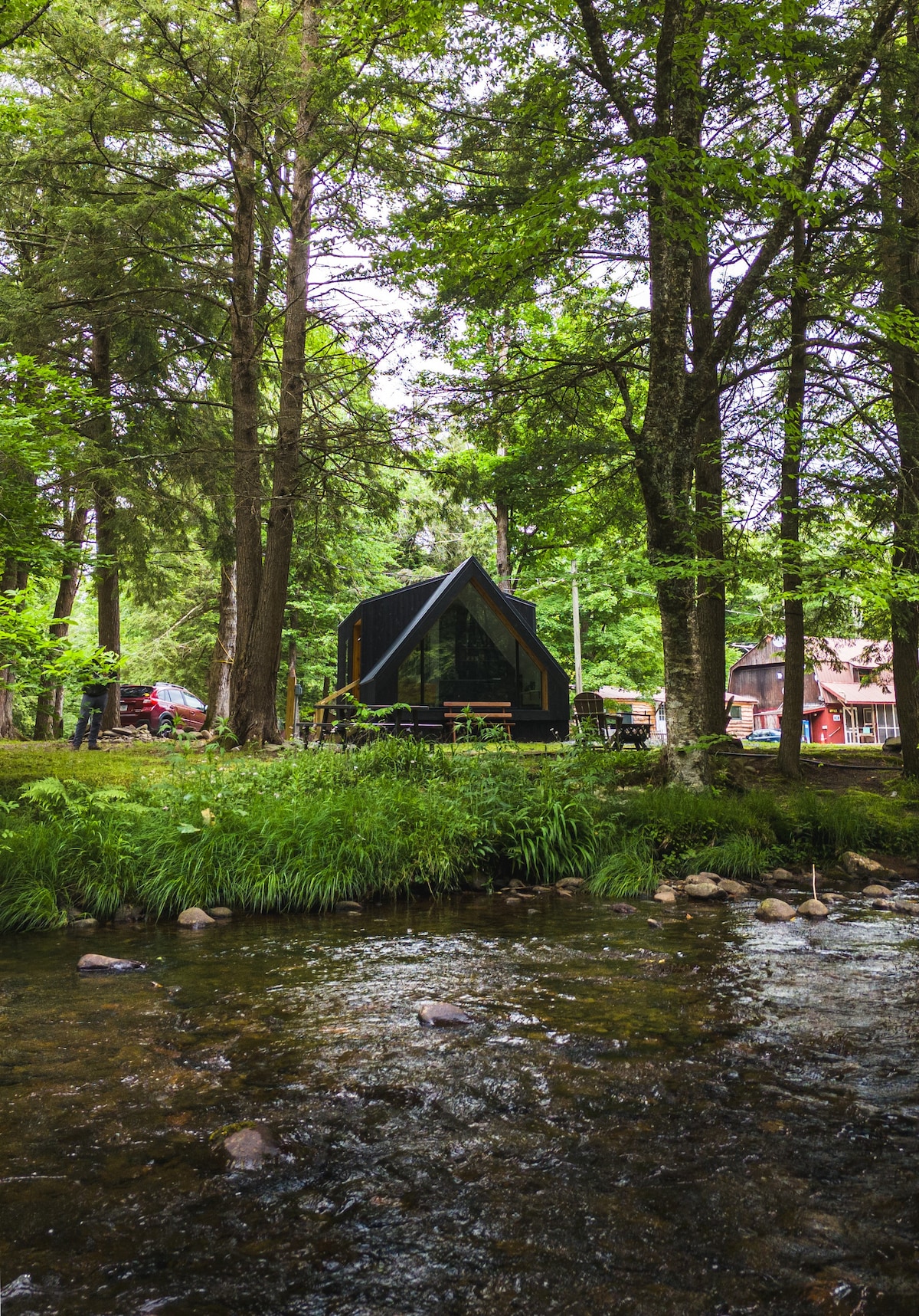 A modern A-frame cabin is set amidst lush greenery beside a gently flowing creek. Trees provide a natural canopy, while rustic outdoor seating can be seen on the deck. The tranquil scene captures the essence of a secluded retreat in a serene natural setting.