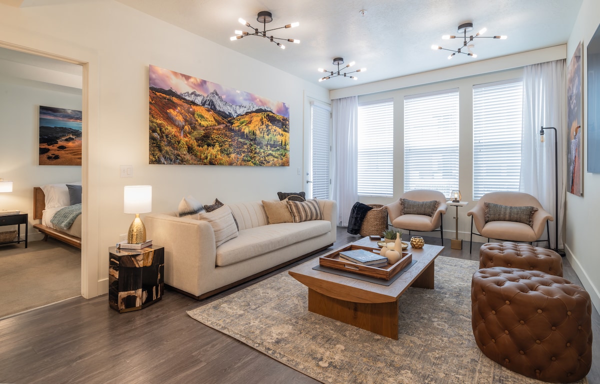 A spacious living room features a light-colored sofa accompanied by two stylish chairs. Natural light fills the room through large windows, with a soft area rug beneath a wooden coffee table. Two artworks depicting scenic landscapes hang on the walls, enhancing the artistic ambience.