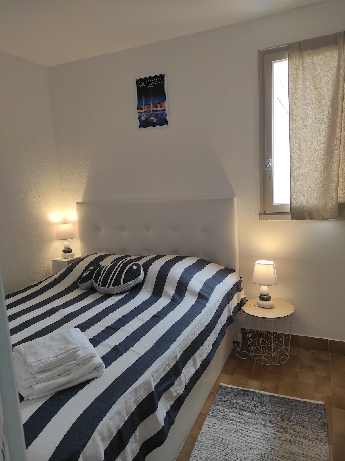 A bright bedroom features a spacious bed adorned with striped bedding and a round accent pillow. Two bedside lamps provide soft lighting, and a window with light drapes allows natural light to enter. A rolled towel is placed at the foot of the bed, adding to the inviting setup.
