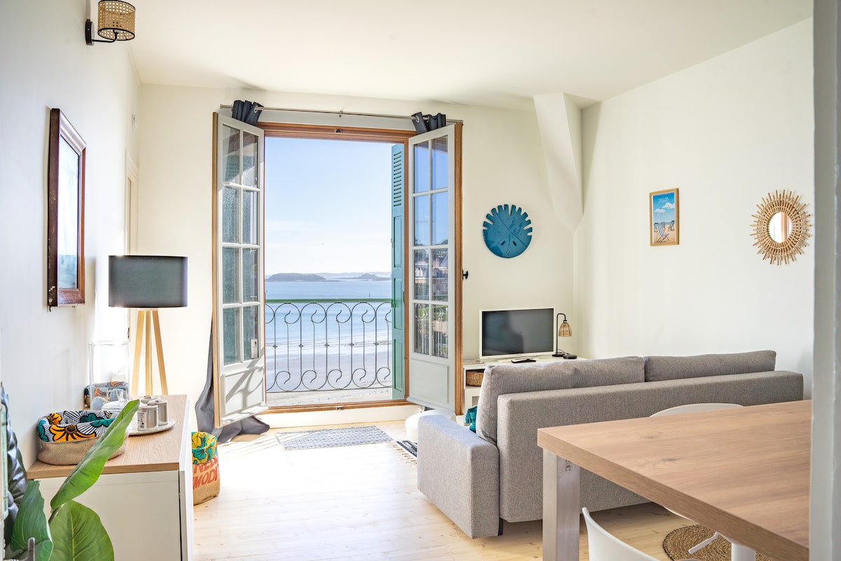 A bright living area is presented with large open windows, offering a scenic view of the sea. A comfortable grey sofa occupies the space, paired with a light wooden dining table. Decorative elements, including wall art and a standing lamp, enhance the inviting environment.