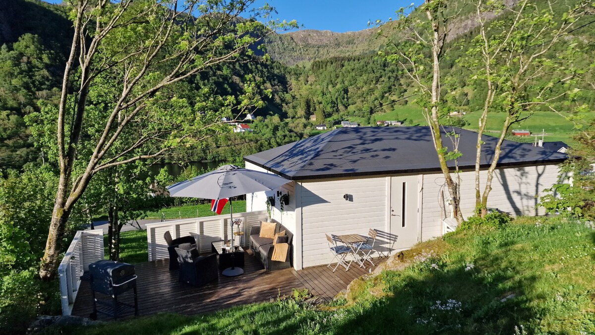 The cabin sits among lush greenery, featuring a spacious terrace equipped with outdoor furniture and an umbrella. The natural landscape forms a scenic backdrop, with hills visible in the distance. A charcoal grill is also present on the terrace for outdoor cooking.