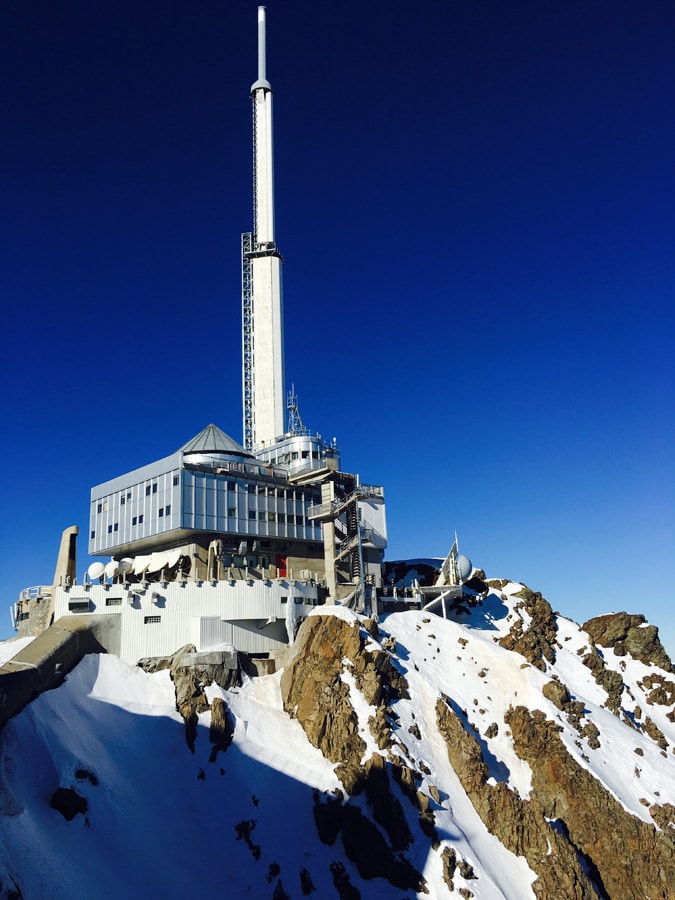 A towering mountain structure with a prominent antenna stands atop a rocky outcrop, surrounded by fresh snow. The clear blue sky contrasts sharply against the building's modern design, showcasing large windows and a angular roofline.