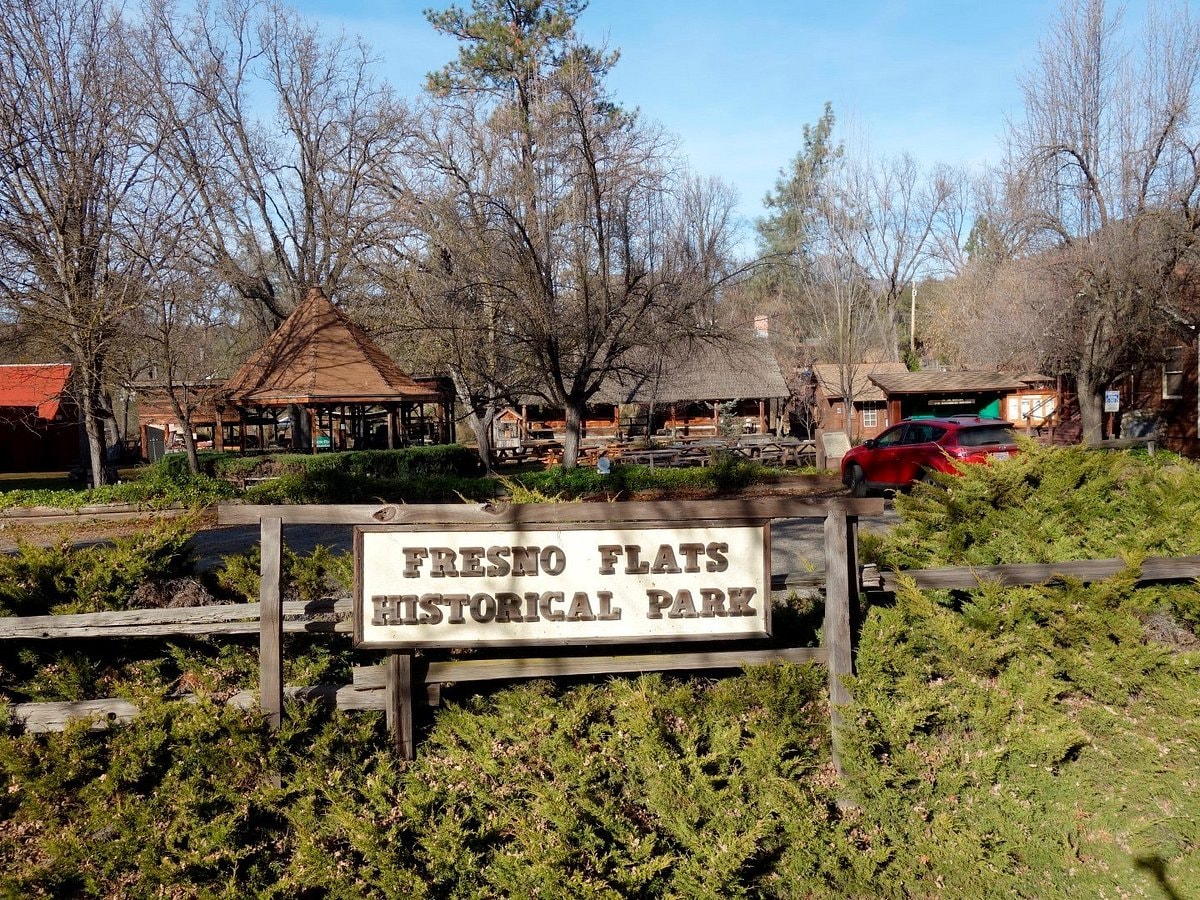 The entrance to Fresno Flats Historical Park is marked by a wooden sign, surrounded by a low wooden fence and lush greenery. In the background, various historic structures are visible, showcasing rustic architecture under a clear blue sky.