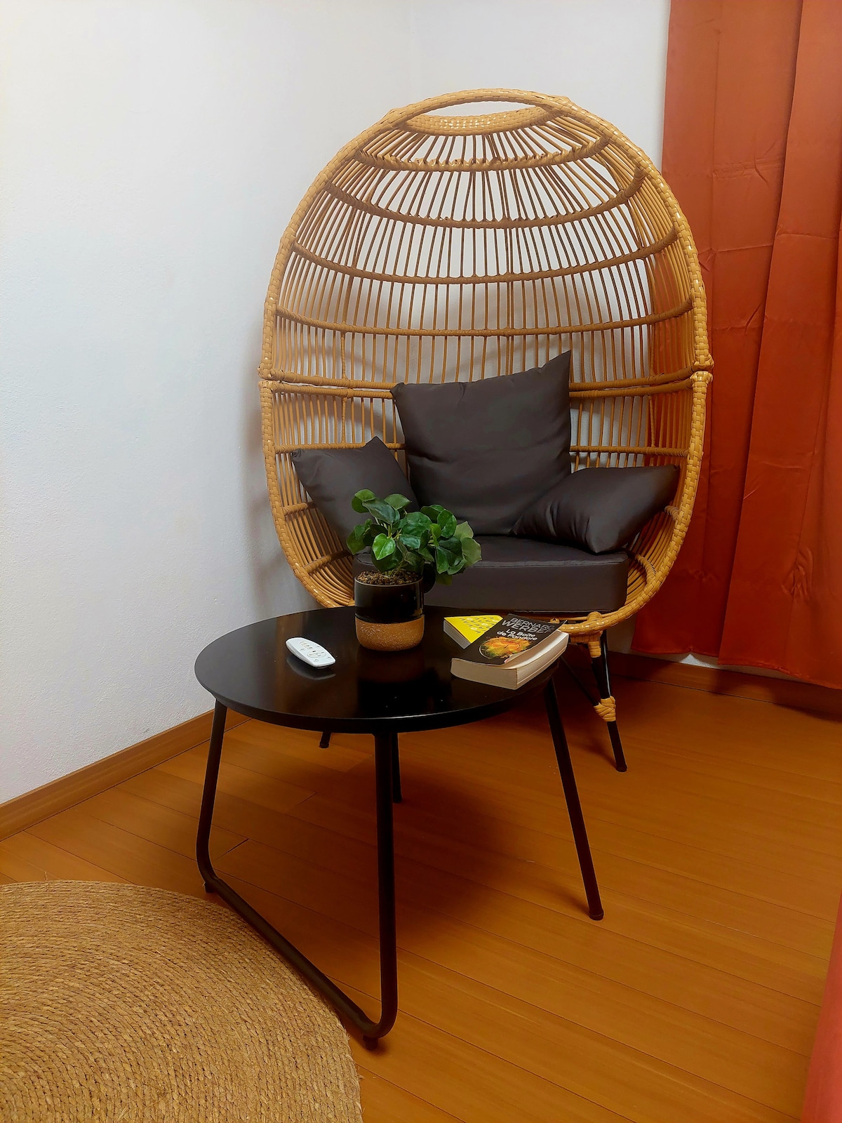 A unique rattan chair with plush cushions is positioned next to a small black table. A potted plant rests on the table, accompanied by a few stacked books. Warm orange curtains frame the space, enhancing the natural tones of the wooden floor.