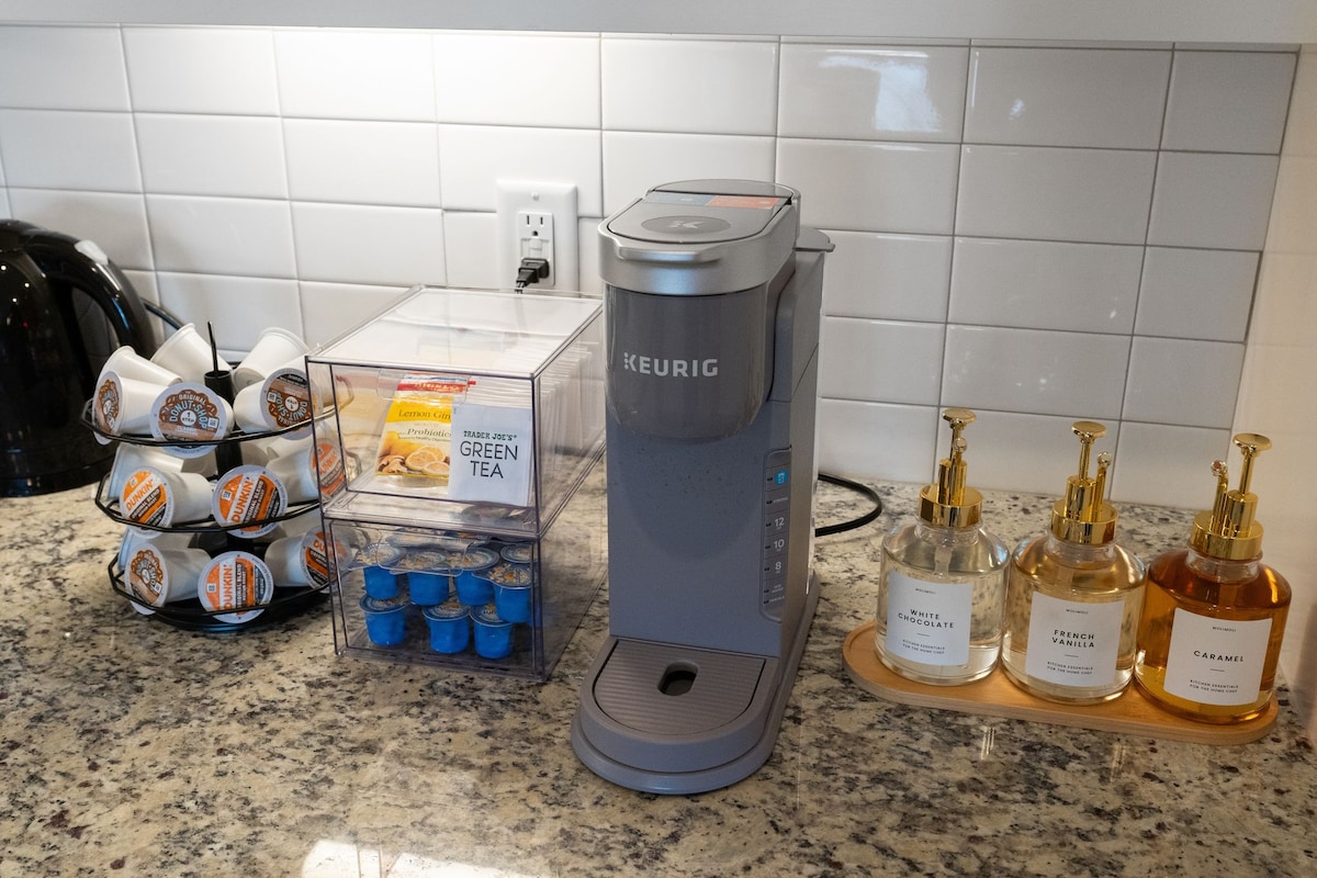 A modern kitchen countertop displays a Keurig coffee maker alongside a container holding assorted coffee pods and green tea. A trio of glass soap dispensers stands on a wooden tray, featuring labels for various personal care products. The surface is topped with a light-colored granite finish.