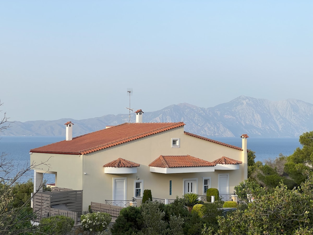 The exterior of a spacious home features a red-tiled roof and light-colored walls, set against a backdrop of mountains and the sea. Lush greenery surrounds the house, adding to the tranquil atmosphere of the property.