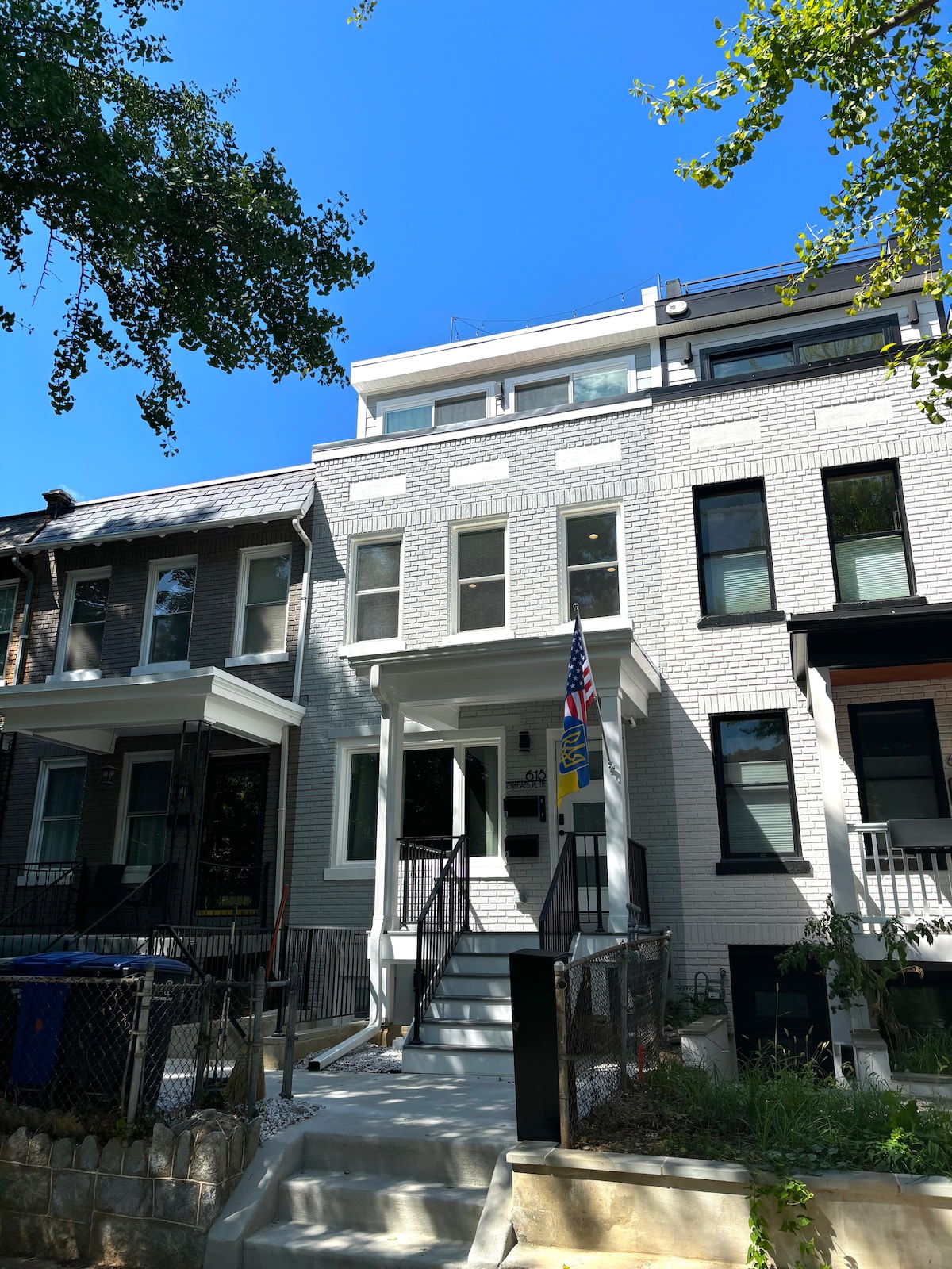 A modern duplex building is presented, featuring a light-colored facade. The structure consists of multiple levels, with a welcoming entrance and steps leading to the front door. Lush greenery surrounds the property, and a clear blue sky is visible above.