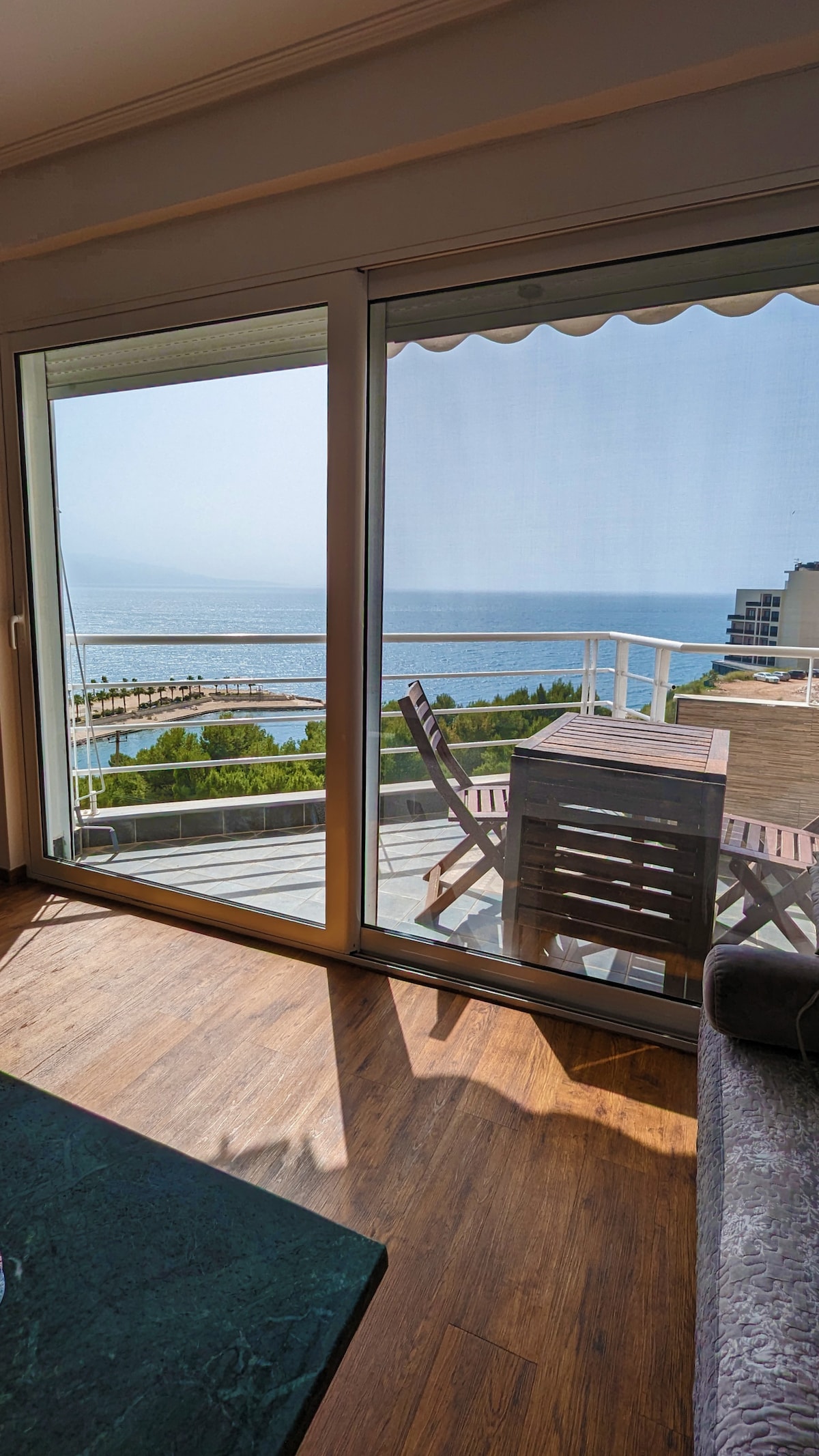 A glass sliding door opens to a spacious balcony with a table and chairs, offering sweeping views of the sea. The wooden floor leads into the living area, where natural light fills the space, enhancing the coastal ambiance.