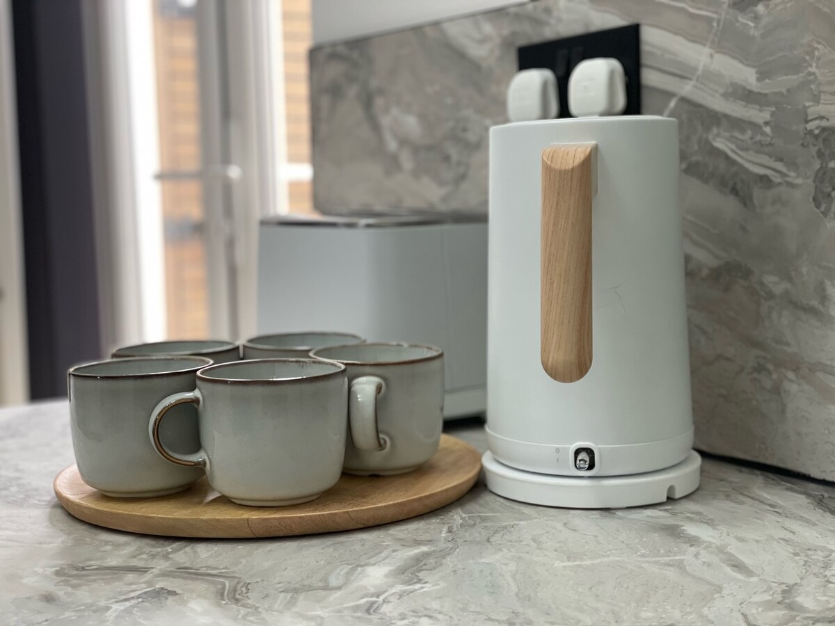 A modern kitchen counter displays a white kettle with a wooden handle alongside a set of four ceramic mugs. The mugs have a subtle grey hue and are arranged on a round wooden tray, creating an organized and functional space.