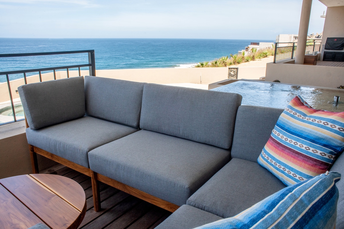 An outdoor seating area features a spacious gray sectional sofa adorned with colorful pillows. A wooden table is nearby, with the coastline visible in the background. The sparkling water of the hot tub is seen on the left, alongside a clear view of the ocean.