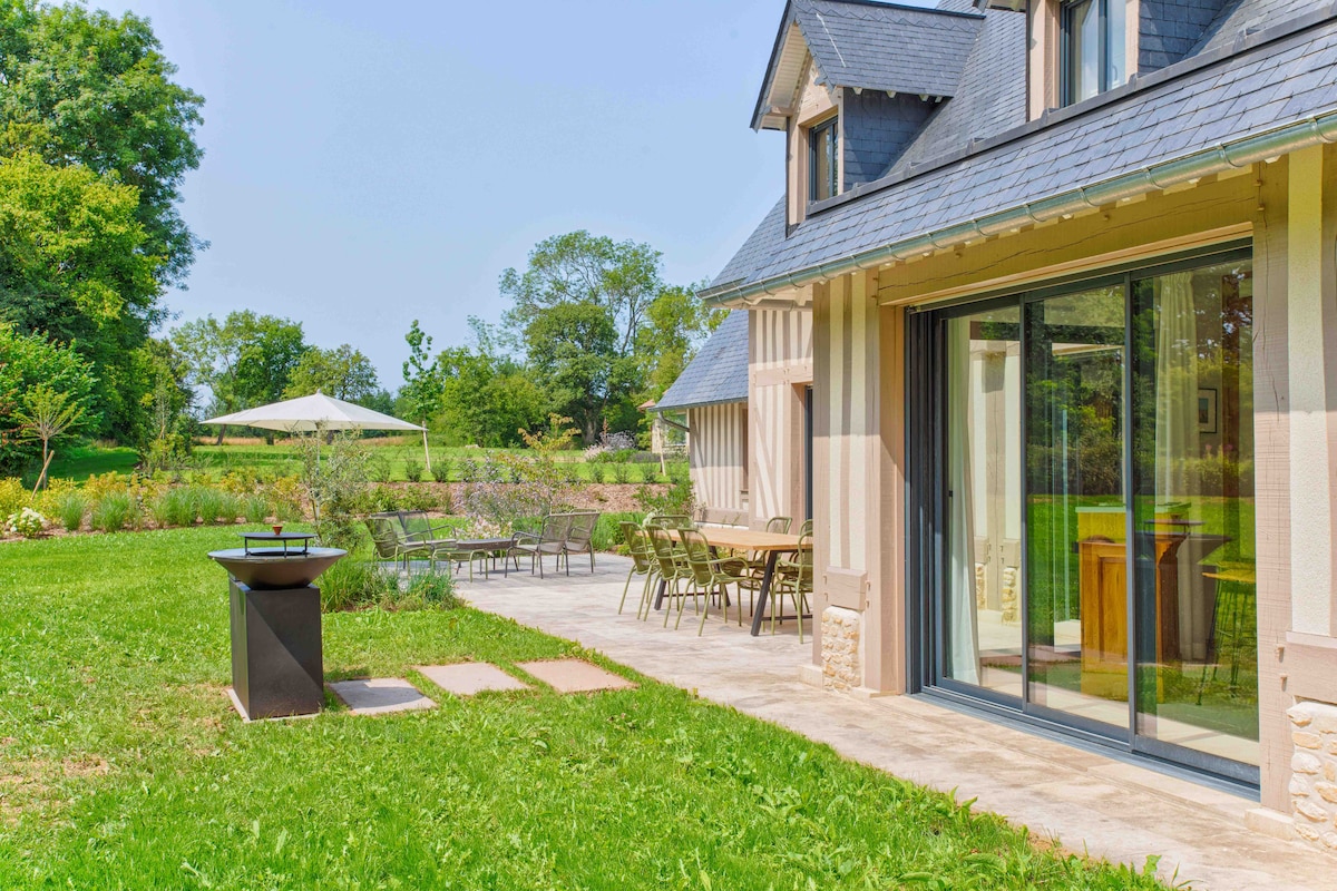 An outdoor terrace features a dining table and chairs, providing ample seating for al fresco meals. A barbecue grill is positioned nearby, while the surrounding grass is complemented by landscaped gardens. Large glass doors connect the terrace to the villa's interior, enhancing accessibility.