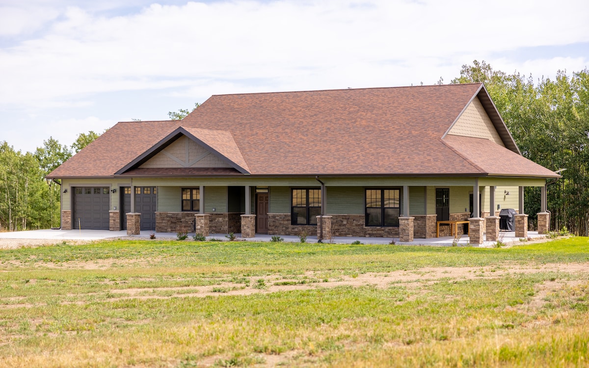 A custom-built ranch-style home is set against a backdrop of lush greenery and an expansive sky. The exterior features a mix of neutral-colored siding and stone accents, with a spacious porch complemented by multiple windows that invite natural light.