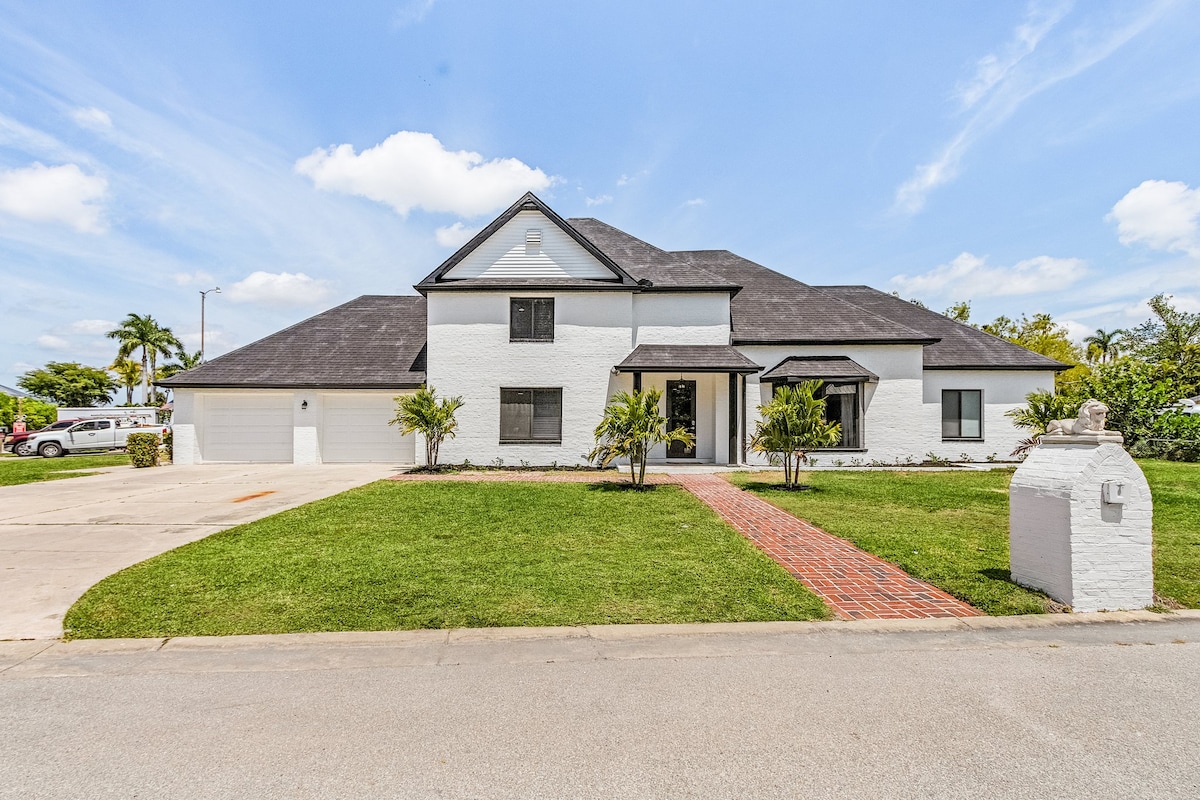 The exterior of a two-story home is presented, featuring a white facade and a dark roof. A landscaped front yard is visible, with green grass and two small palm trees flanking the entrance. A brick pathway leads to the front door, while a spacious driveway accommodates multiple vehicles.