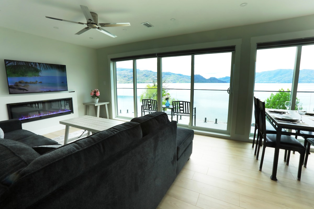 A spacious living area features large windows offering expansive lake views. Comfortable seating is arranged around a modern coffee table. A dining table prepared for four is visible, while a minimalist entertainment setup includes a flat-screen TV and a contemporary electric fireplace.