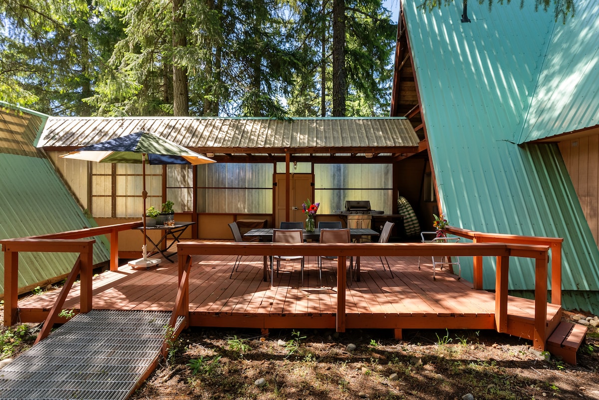 A welcoming deck is surrounded by lush trees, featuring a round table with comfortable chairs and an umbrella for shade. The unique A-Frame cabin design showcases angled roofs and large windows, creating a connection to nature while providing an inviting space for outdoor gatherings.