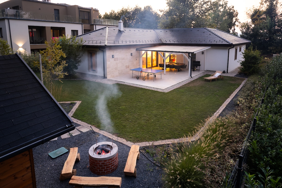 An outdoor space features a landscaped garden with green grass and a stone fire pit surrounded by wooden benches. A ping-pong table sits on a spacious terrace, while large glass doors allow views into the house. Subtle smoke rises from the fire pit as evening approaches.