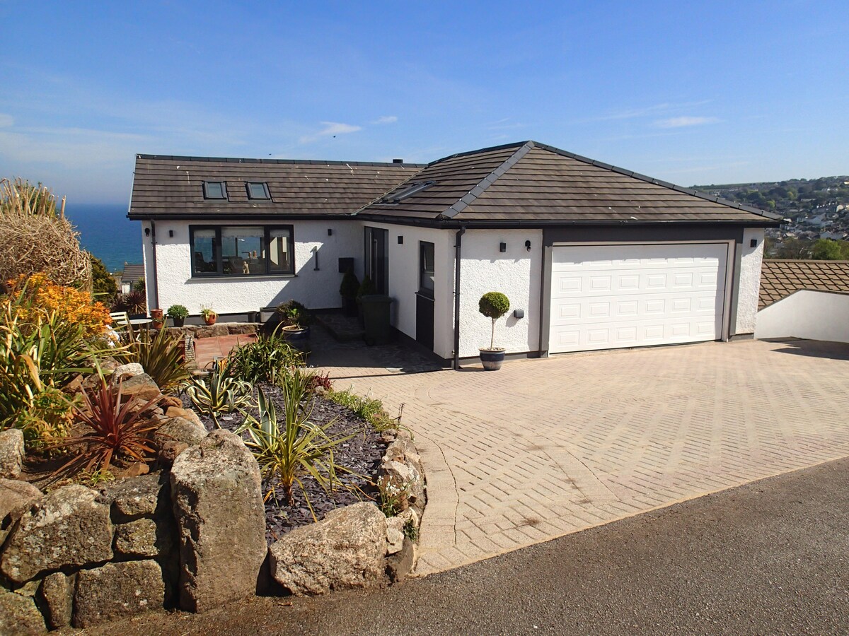A modern coastal home is shown, featuring a smooth, paved driveway that leads up to a double garage. Lush plants and low stone walls enhance the exterior, with a clear view of the sea visible in the background under a blue sky.