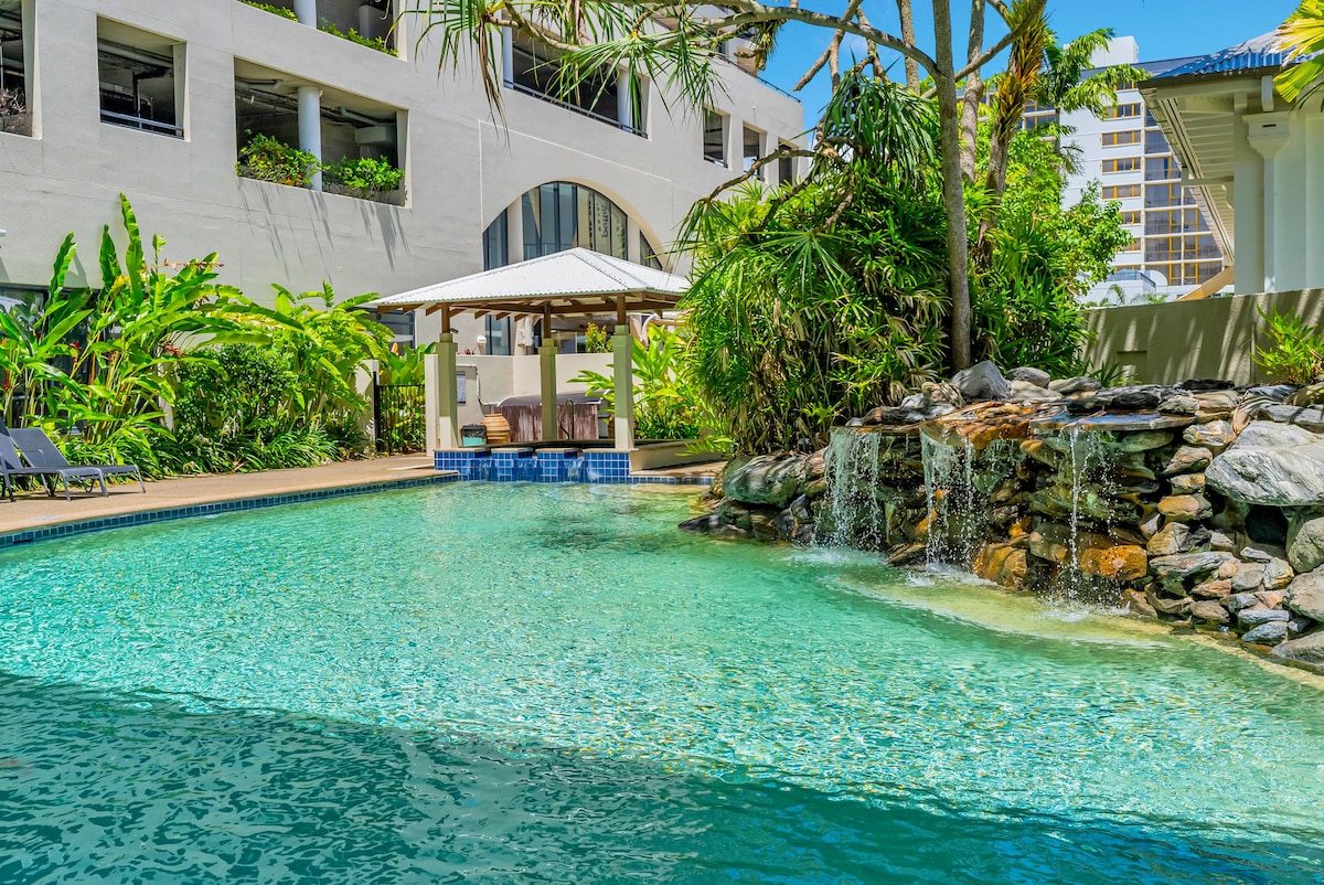 A serene swimming pool is surrounded by lush tropical landscaping, featuring a rock waterfall that gently cascades into the clear water. A shaded gazebo with seating is visible nearby, offering a relaxing space for guests to enjoy the tranquil environment.