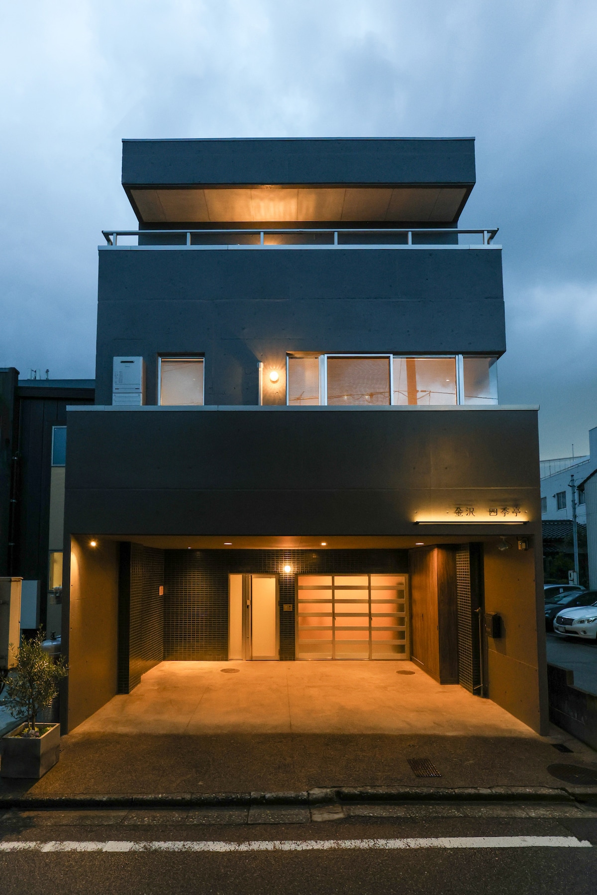 A three-story building features a modern façade with large windows, allowing for ample natural light. The entrance is highlighted by warm exterior lighting, and a spacious parking area is visible in front of the structure.