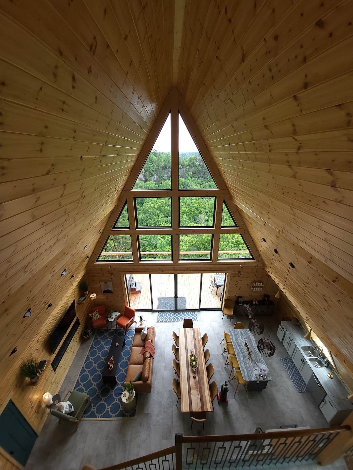 An aerial view of a modern A-frame interior highlights soaring wooden ceilings and expansive windows, allowing natural light to fill the space. A long dining table is positioned centrally, surrounded by multiple chairs. A cozy seating area is arranged to one side, with soft furnishings.