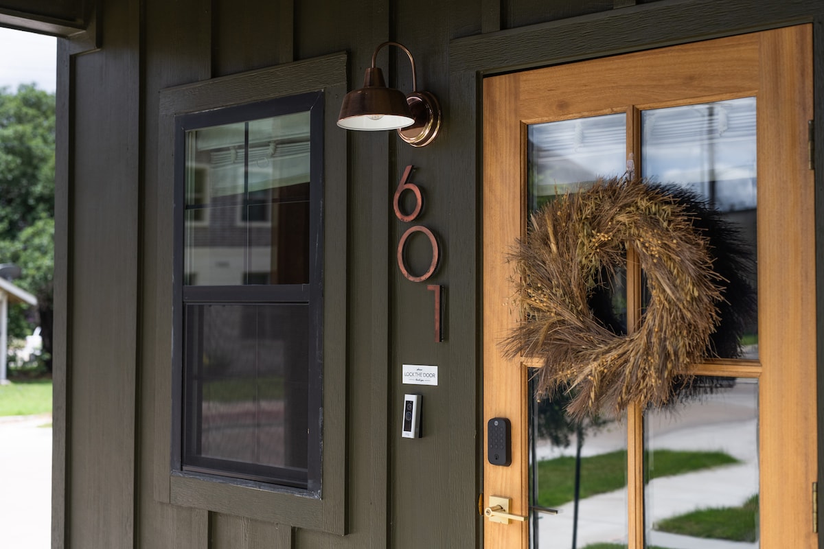 The entrance features a sturdy wooden door adorned with a natural wreath. An elegant light fixture is mounted above, and the house number is displayed prominently. A window beside the door allows for natural light, with a view of the landscaped exterior.