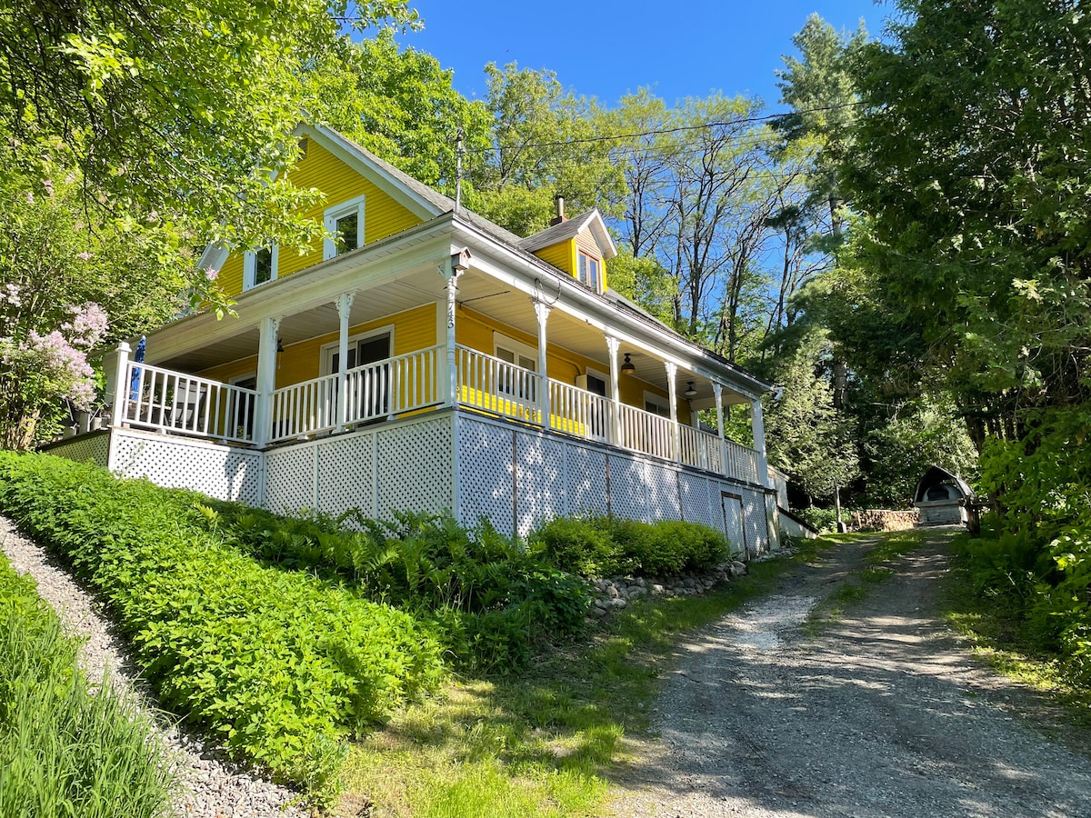 A charming yellow chalet is nestled among lush greenery, featuring a welcoming front porch with white railings. The gravel driveway leads to the entrance, emphasizing the peaceful natural surroundings with trees overhead.