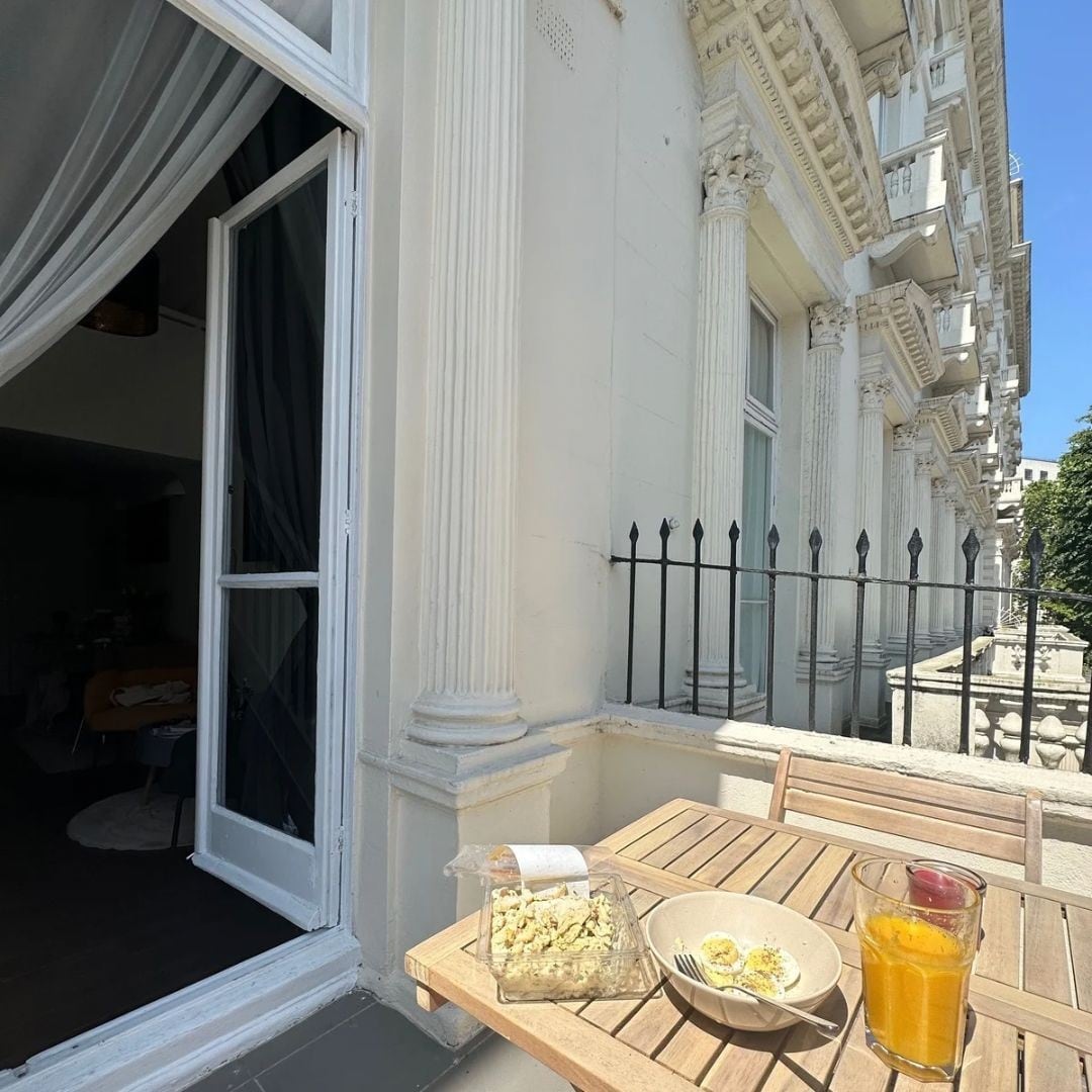 A wooden table set on a balcony is adorned with a bowl of popcorn and a glass of orange juice. The elegant exterior features classic architectural details, while sheer curtains softly frame the open doorway, allowing light to enter from the living space inside.