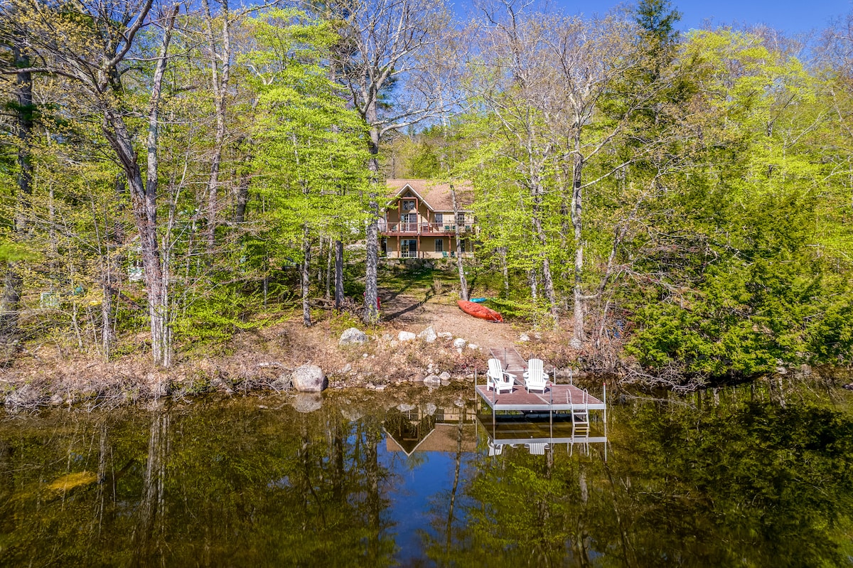 A serene lakeside view features a wooden home nestled among lush trees. A private dock with two chairs and a red kayak is positioned on the calm water, reflecting the vibrant greenery and the structure above.