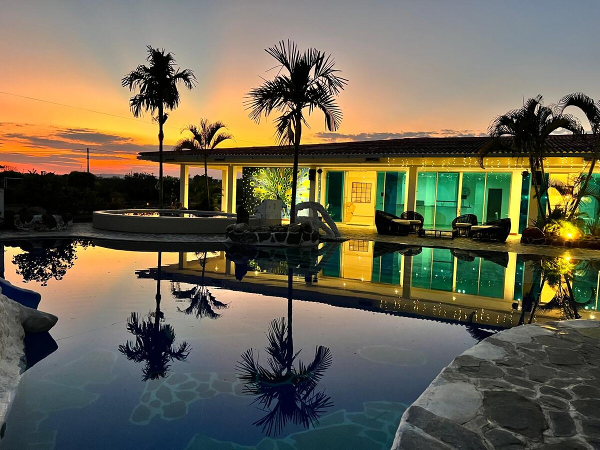 A tranquil swimming pool reflects the vibrant hues of a sunset, surrounded by lush palm trees. Soft lighting from the nearby building casts a warm glow on the water's surface, creating a calming and inviting atmosphere.