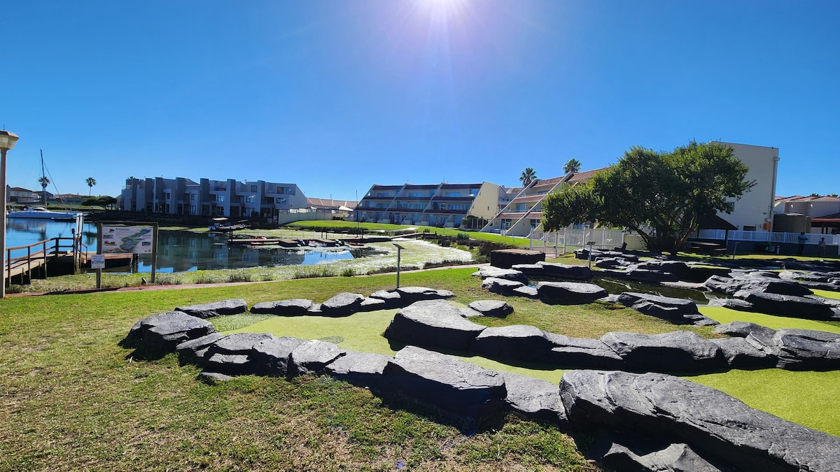 A landscaped area features smooth black stones arranged across lush green grass, creating a striking contrast. The serene waterway reflects the clear blue sky, while several buildings can be seen in the background, adding context to the tranquil setting.