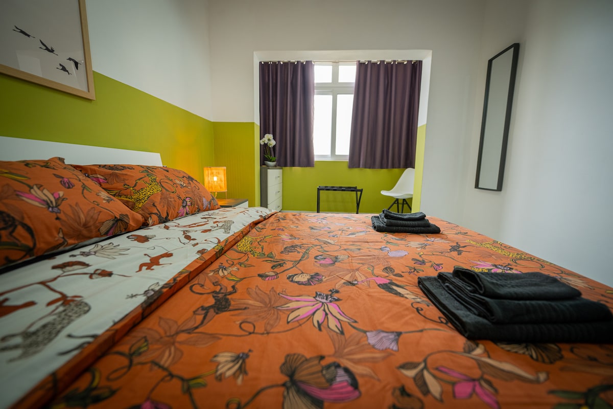 A brightly decorated bedroom features a spacious bed adorned with floral-patterned bedding in warm orange hues. A large window allows natural light to fill the room, and rich purple curtains frame the view. A small nightstand and a chair complete the inviting space.